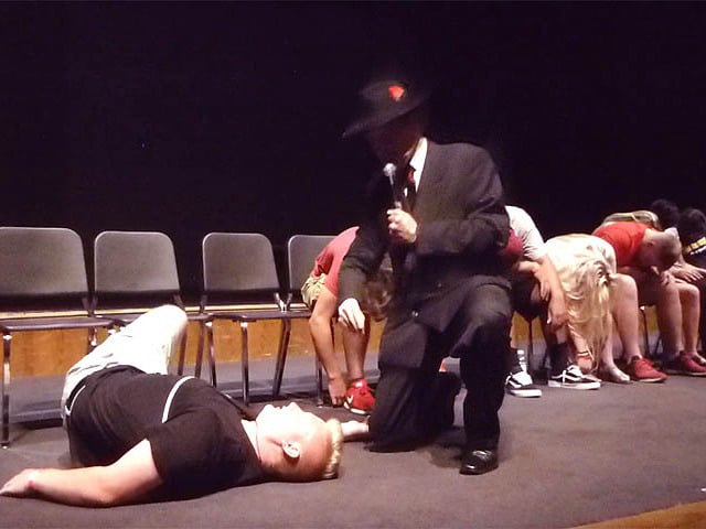 Hypnotist Mister Vibe speaks to a male student on the floor at a student falls out of his seat at a Chicago area high school graduation hypnosis show.